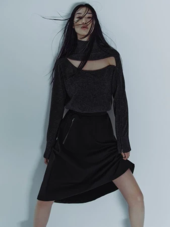 A model with long dark hair blowing across her face wearing a dark cut-out top and black skirt against a pale wall.