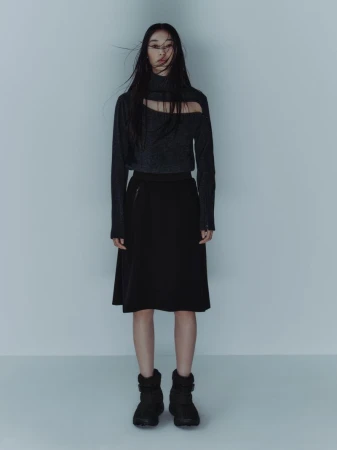 Model with long dark hair wearing a dark cut-out sweater and black skirt against a pale background