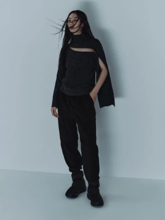 Model with long dark hair blowing in the wind, wearing a cut-out grey sweater and black trousers against a neutral background.