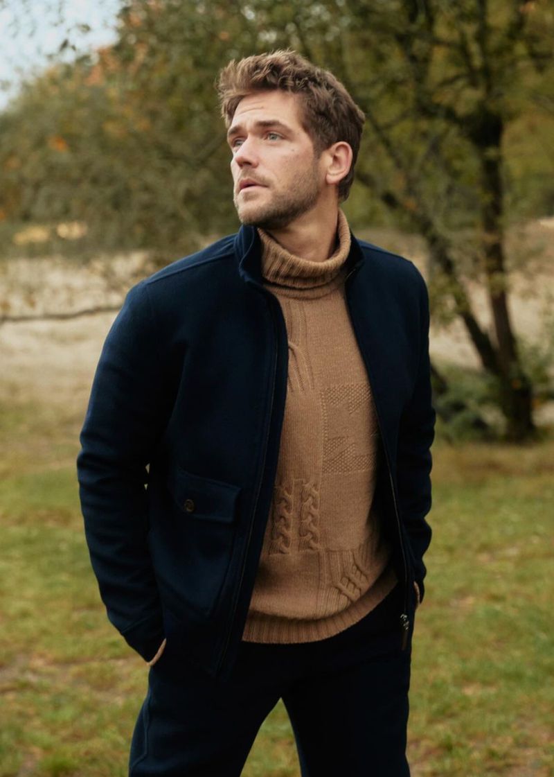 Handsome man in a navy jacket and tan turtleneck sweater stands outdoors against a green and brown foliage background, looking away.