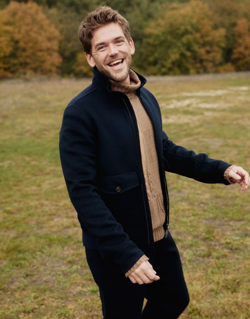 Smiling adult man in navy jacket and brown sweater, standing outdoors in field, captured at eye-level, with bokeh effect and natural light.