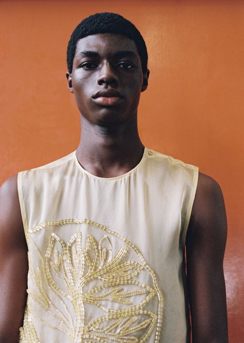 Portrait of a young adult male with short dark hair wearing a semi-sheer sleeveless shirt with beaded floral design in front of an orange wall.