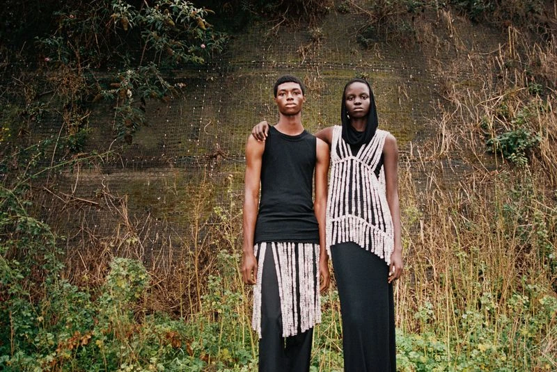 An adult male and female couple pose outdoors against a textured wall and natural foliage, both in black clothing with neutral-toned beaded garments.