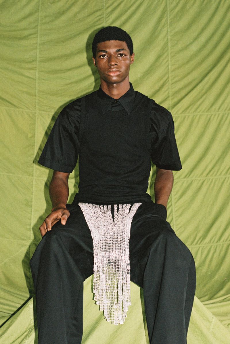 Man in black shirt and pants seated against a green backdrop with a decorative net-like apron-style garment.