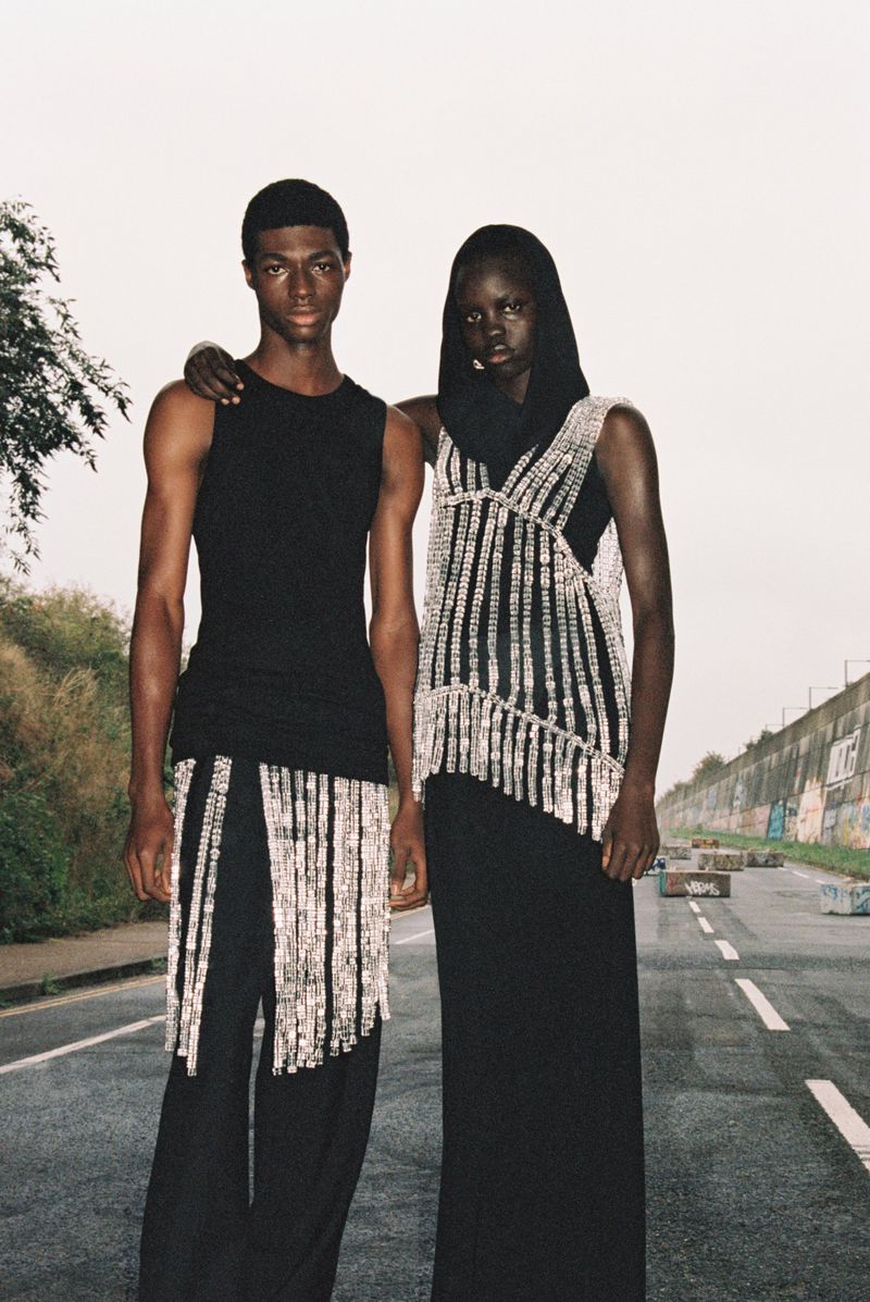Two fashion models in black outfits pose on a road with light sky. The model on the right wears a silver beaded top with fringe.