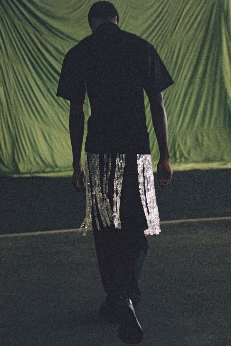 Adult male fashion model walks away, wearing a black t-shirt, pants and silver fringe around his waist. Green draped background.