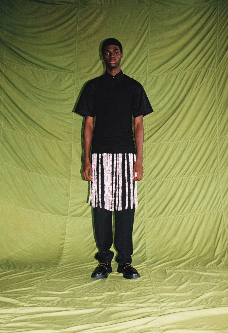 A young adult Black male stands in a studio wearing a black polo shirt, patterned short skirt, black pants, and black shoes, against a green backdrop.