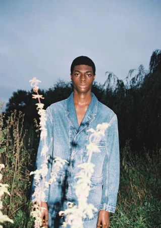 Portrait of a young adult Black man wearing a blue denim jacket in an outdoor setting with wild plants in the foreground.