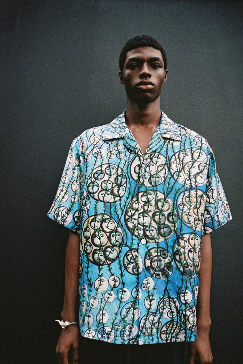 Portrait of a young adult male in a patterned shirt, standing against a dark wall. Fashion shot with natural lighting.