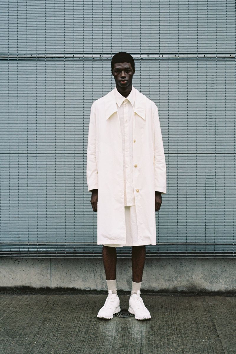 Full body shot of a slender, dark-skinned male model in a white trench coat, shorts, and sneakers against a textured blue wall.