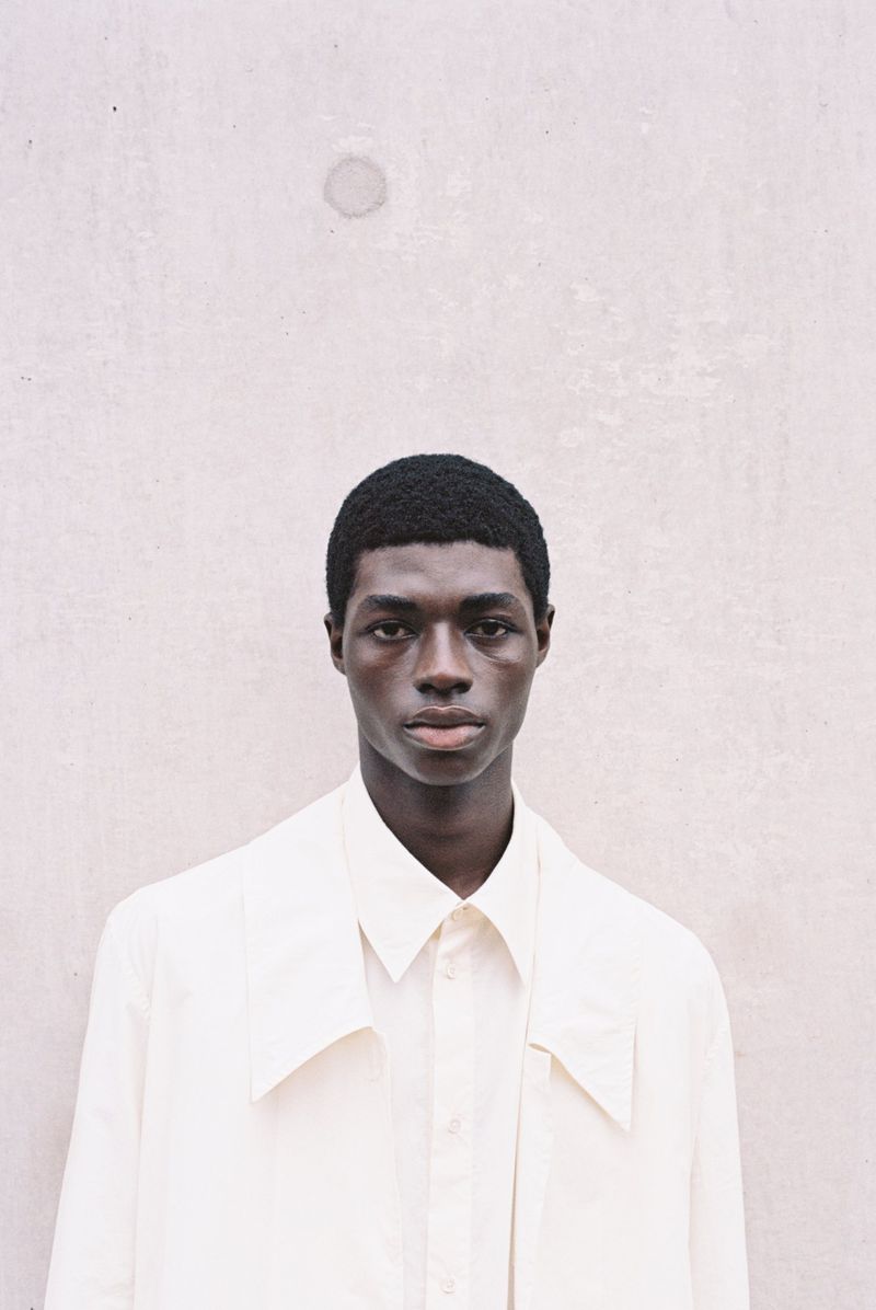 Close-up portrait of a Black male model in a white shirt and jacket against a neutral wall, showcasing fashion and minimalist style.