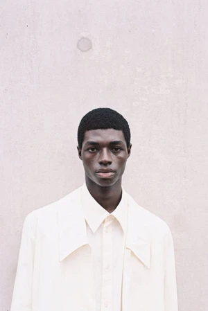 Portrait of a black male model wearing an off-white collared shirt against a textured concrete wall.