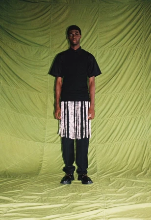 Model standing against a quilted olive green fabric backdrop wearing a black polo shirt and fringe skirt over trousers