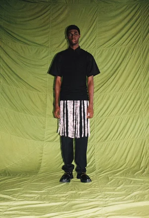 Model standing against a quilted olive green fabric backdrop wearing a black polo shirt and fringe skirt over trousers
