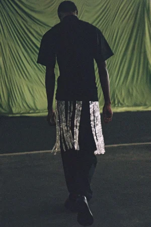 Model Zaram Obasi seen from behind wearing black shirt and trousers with beaded skirt overlay against draped green background.