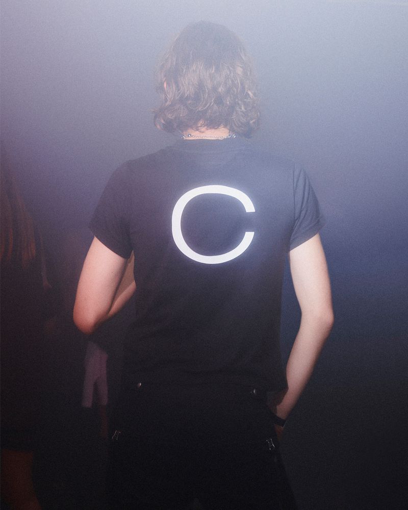 Person with medium-length wavy brown hair faces away, wearing a black shirt with a large white 'C' on the back in a hazy environment.