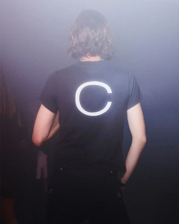 Person with medium-length wavy brown hair faces away, wearing a black shirt with a large white 'C' on the back in a hazy environment.