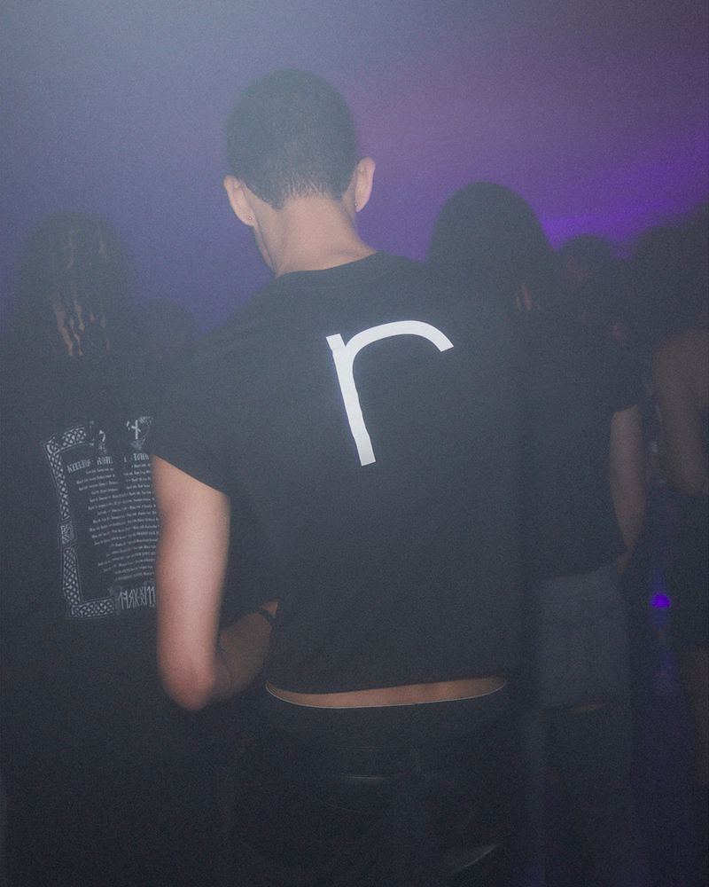Person in a black t-shirt with a white letter 'r' at an indoor event with other people in the background under dim lighting.