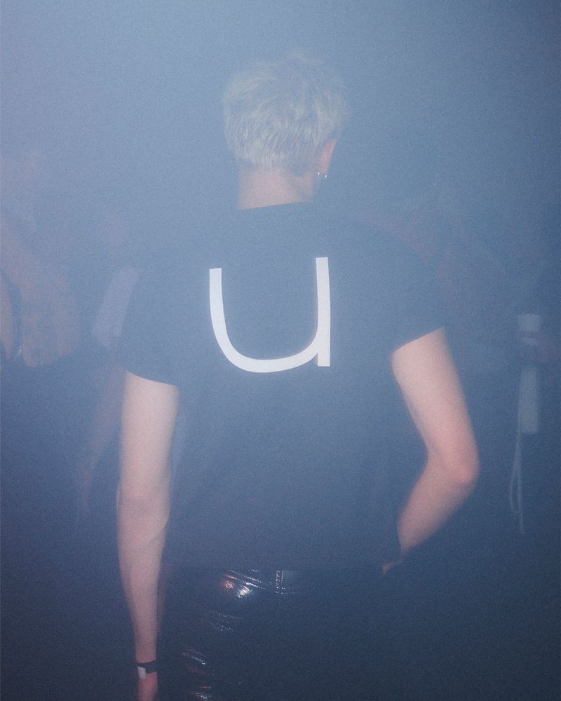 Person in black shirt and shiny black pants indoors in a foggy environment with back to the camera. White 'U' letter on shirt.