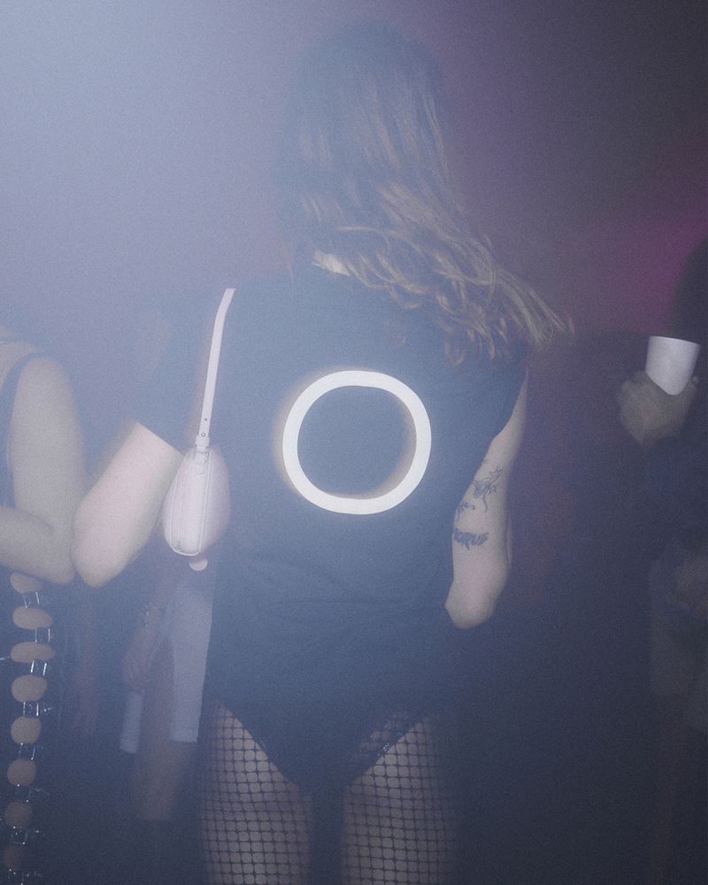 A person in a black shirt with a glowing white circle design on the back, standing in a dimly lit indoor venue with other people present.