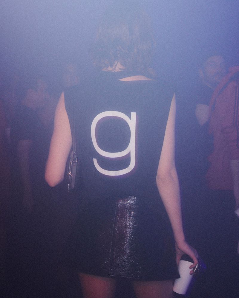Back view of a young adult female with dark hair wearing a black top with a white 'g' and a metallic skirt in a dark, smoky indoor setting.
