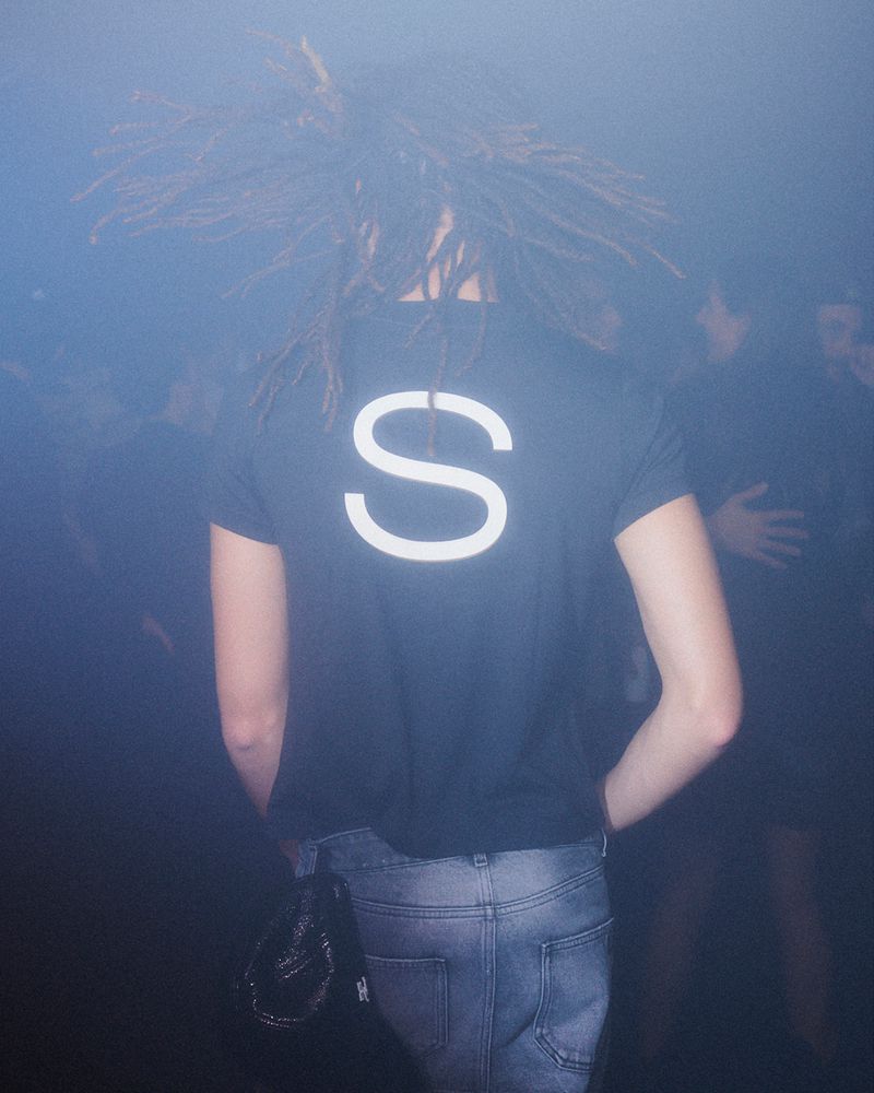 Person with dreadlocks and a black t-shirt with the letter S stands in a smoky indoor environment with other people in the background.