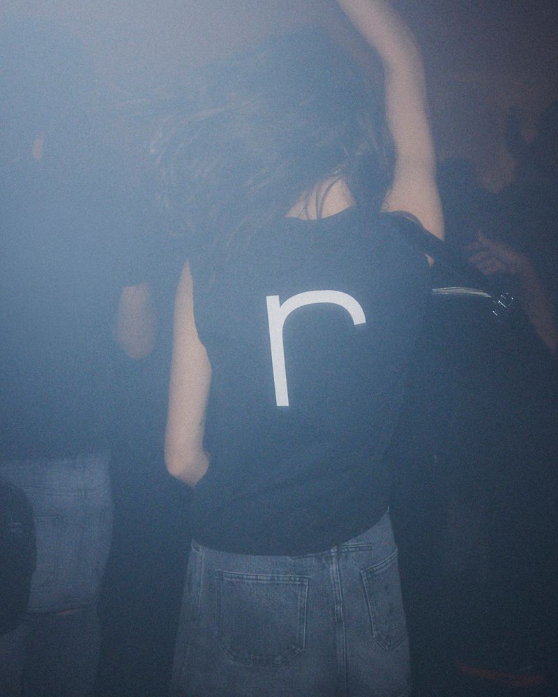 Person in jeans and a black t-shirt with a large white 'r' dancing in a dark, crowded indoor venue with hazy, dark lighting.