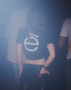 Woman wearing a black logo t-shirt and dark trousers seen from behind in a hazy club setting