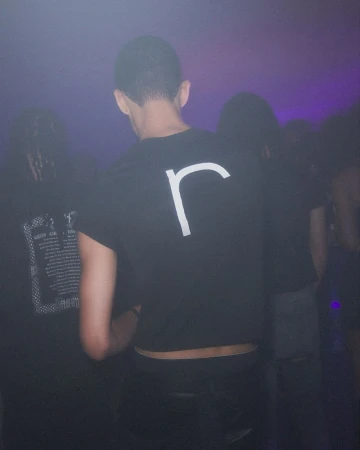 Group of people in a dimly lit club setting, focusing on a person from behind wearing a black t-shirt with a white letter r.
