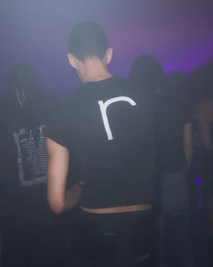 Group of people in a dimly lit club setting, focusing on a person from behind wearing a black t-shirt with a white letter r.