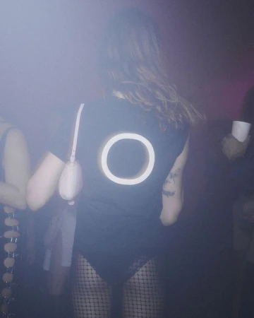 Model wearing a black t-shirt with a glowing circular design on the back and fishnet tights in a hazy nightclub setting