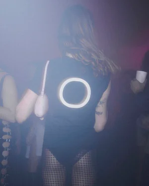 Model wearing a black t-shirt with a glowing circular design on the back and fishnet tights in a hazy nightclub setting