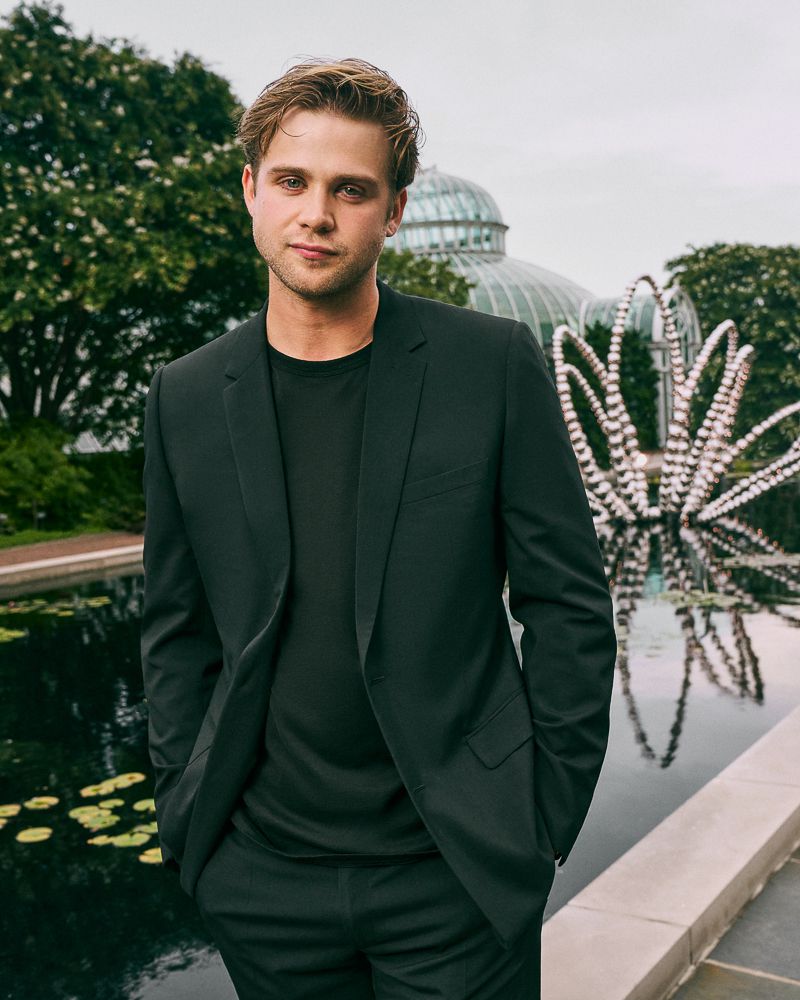 Portrait of an adult male in a black suit in an outdoor setting with greenery, a pond, and a glass building in the background.
