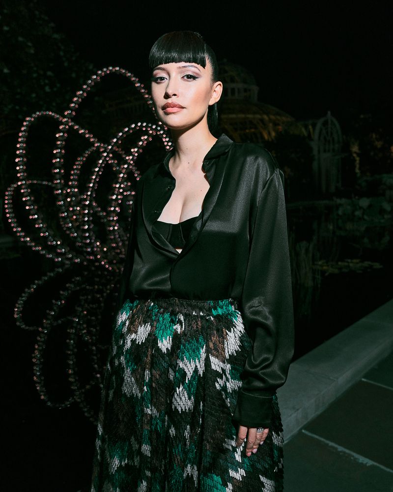 Portrait of a young woman with bangs and a dark silk blouse posing outdoors with an abstract sculpture and dark foliage in the background.