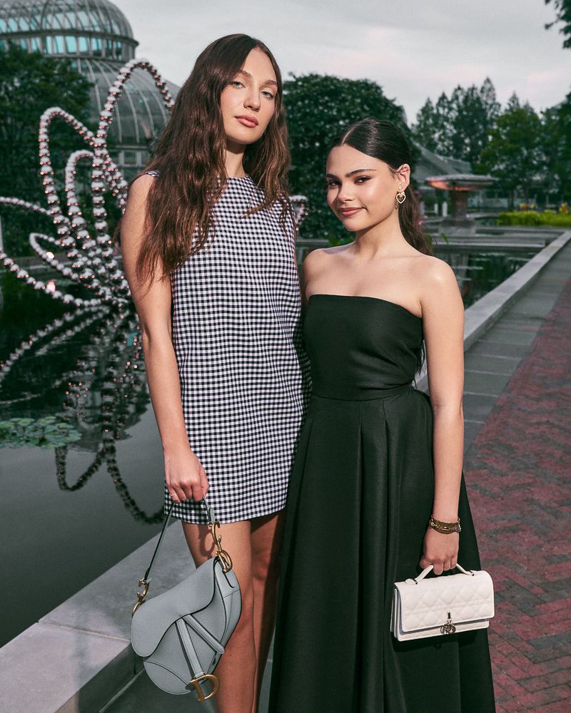 Two women pose in elegant dresses with handbags in an outdoor garden setting with a glass dome structure and pond in the background.