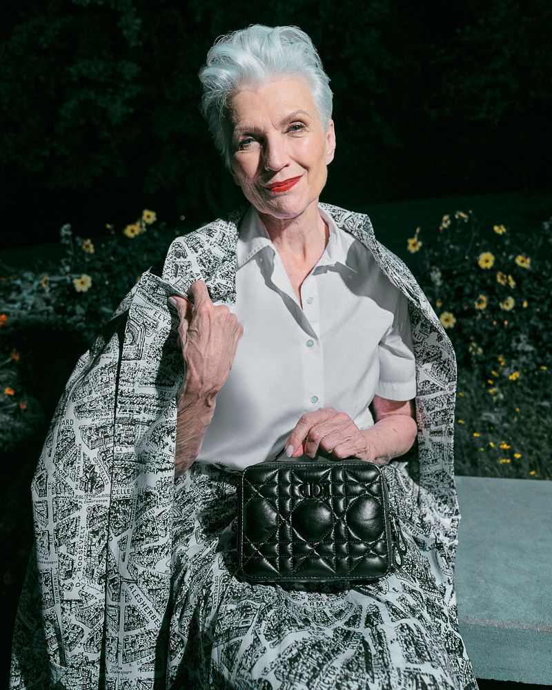 Mature woman with silver hair, wearing white blouse, patterned skirt and jacket, holding a black purse, sitting outdoors in a garden.