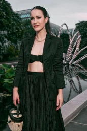 Rachel Brosnahan in a black blazer and skirt holding a small structured handbag outdoors in front of a glass greenhouse structure