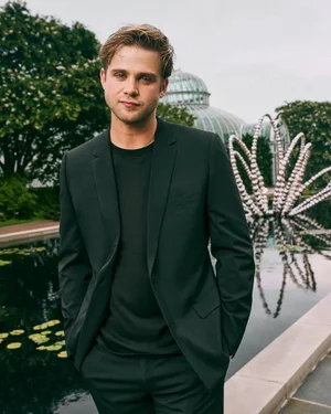 Actor Thomas Doherty wearing a black blazer and t-shirt standing by a reflecting pond with a conservatory behind him.
