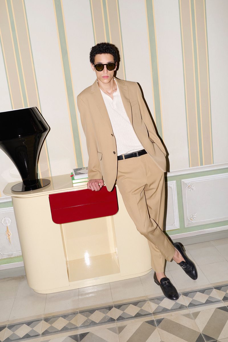 Man in a tan suit poses indoors, leaning against a cream-colored cabinet with a black vase, wearing sunglasses and black loafers.