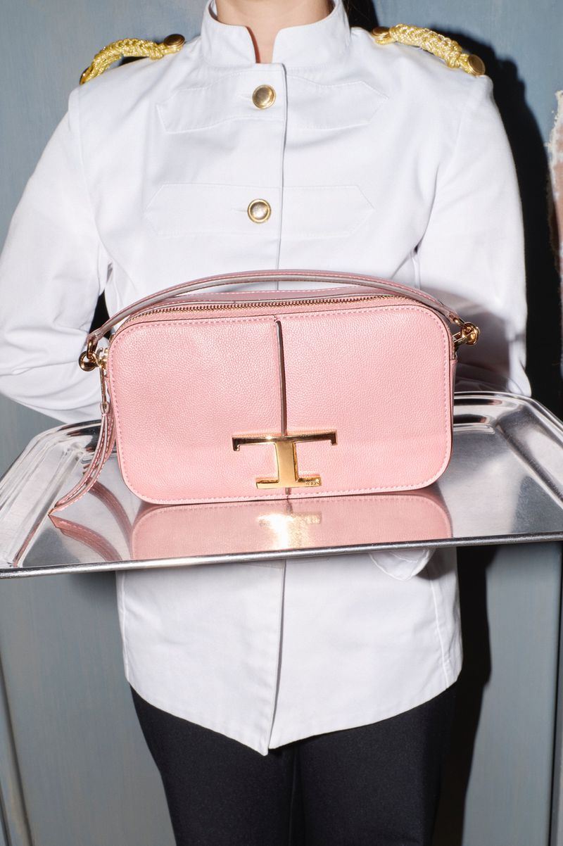 Close-up of a pink leather handbag with a gold logo being held on a silver tray by a person in a white uniform jacket.