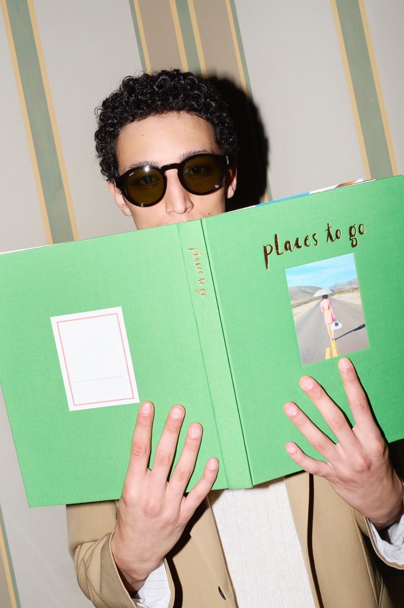 Adult man with sunglasses is holding a green book titled 'Places to go' over his face, showcasing a desert road picture.