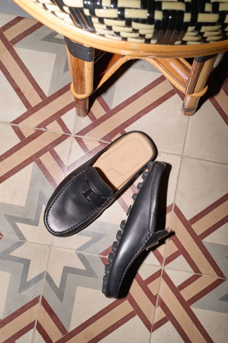 Black leather mule shoes with a stacked heel sit on a patterned tile floor next to a woven chair in a brightly lit indoor space.