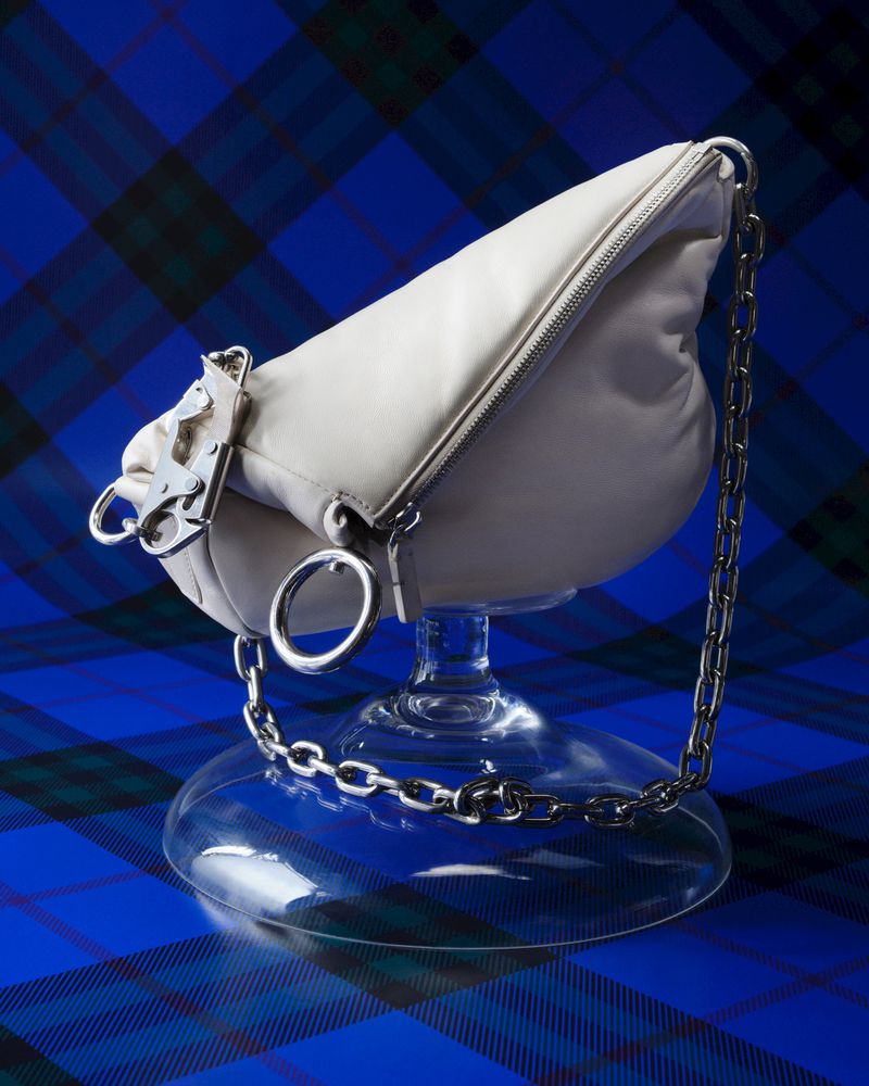 Studio shot of a cream-colored, soft, leather bag with silver hardware, resting on a clear glass stand, against a blue tartan background.