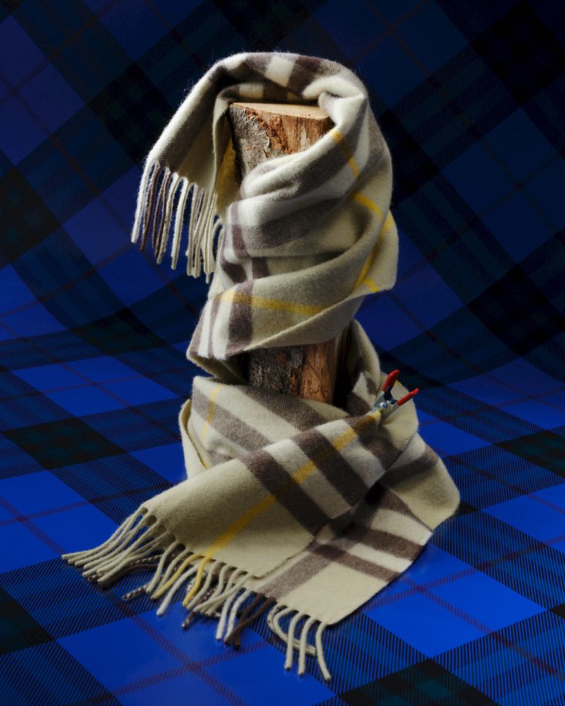 Beige and brown plaid scarf draped on a tree stump against a blue and black plaid textile backdrop, creating a textured indoor scene.