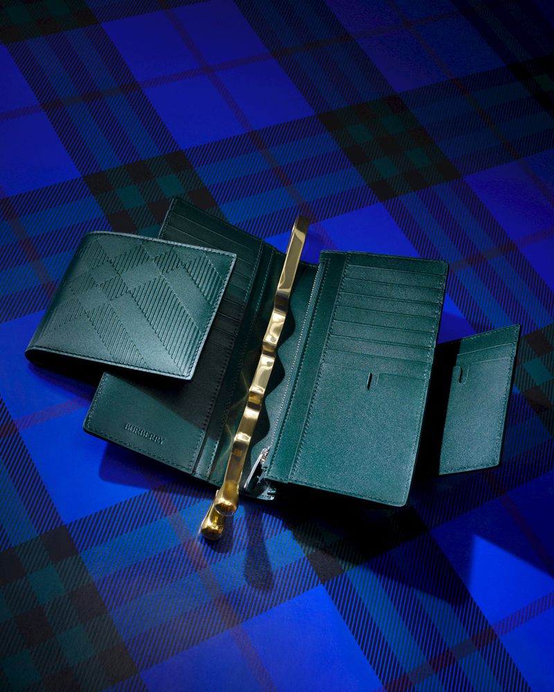 Overhead studio shot of green leather wallets and a gold-colored zipper puller on a blue and black tartan background.