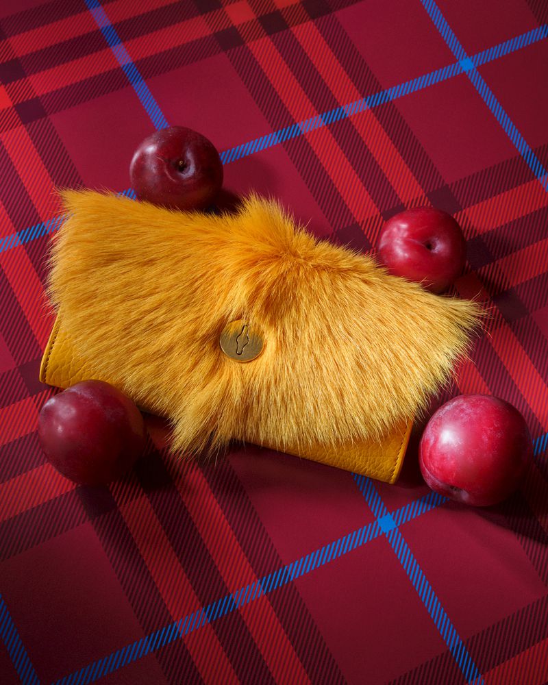 Still life of a yellow fur clutch surrounded by plums on a red and blue plaid surface, fashion accessory, fruit.