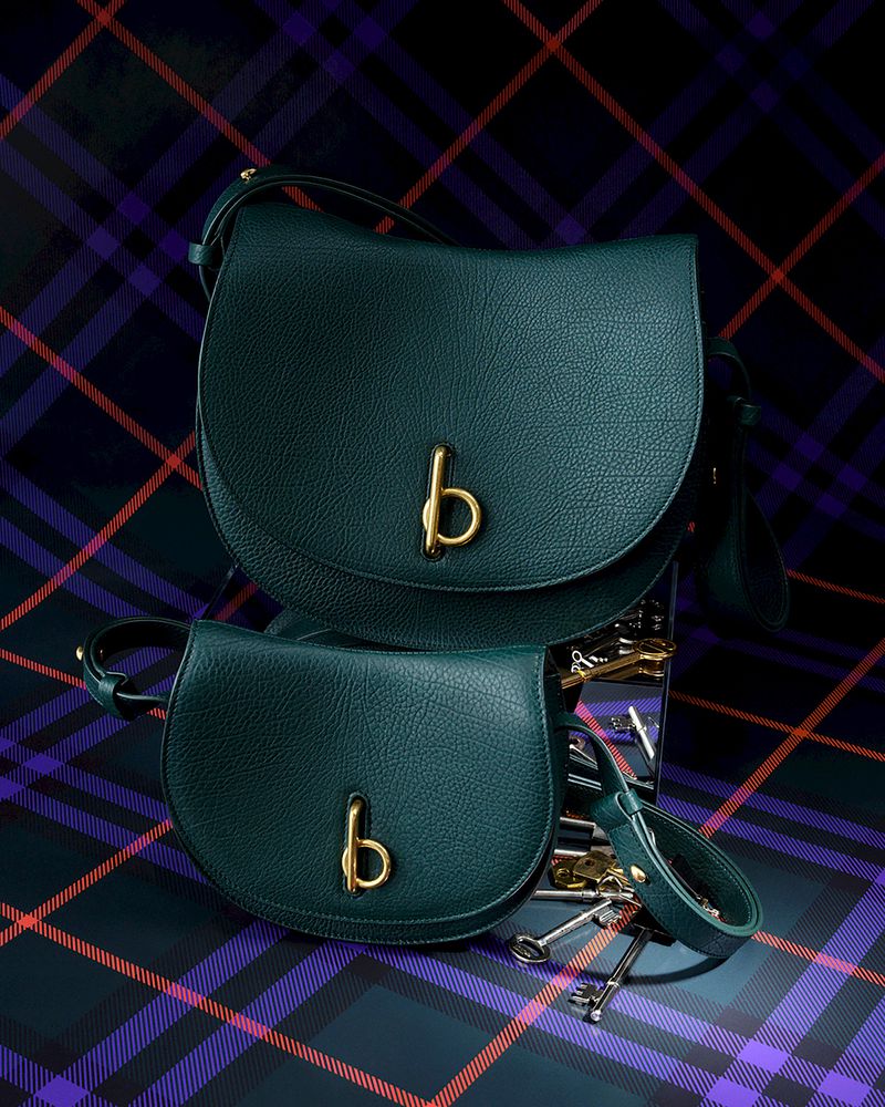 Two green leather saddlebags with gold hardware are displayed on a reflective surface against a blue and red tartan background.