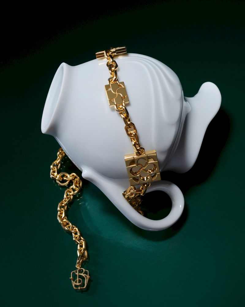 Close-up of a gold chain necklace draped on a white ceramic teapot against a dark green background, showcasing jewelry and tableware.
