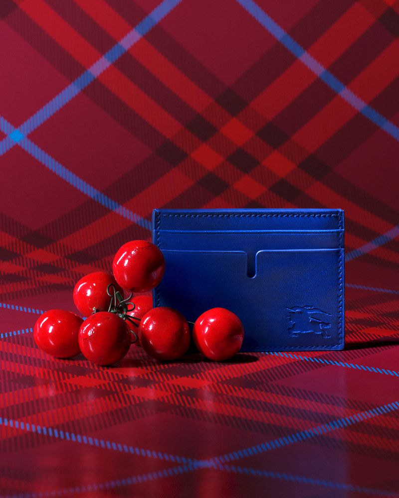 Blue leather card holder rests against a backdrop of red plaid fabric, surrounded by a cluster of vibrant red cherry tomatoes.