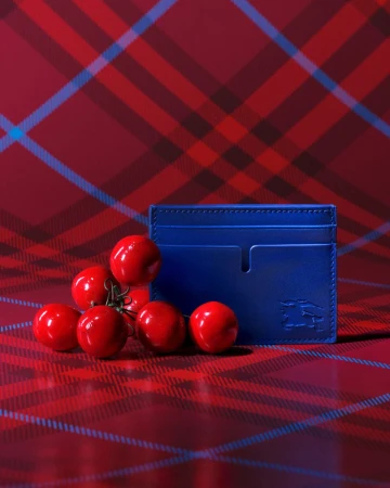 Blue leather card holder rests against a backdrop of red plaid fabric, surrounded by a cluster of vibrant red cherry tomatoes.